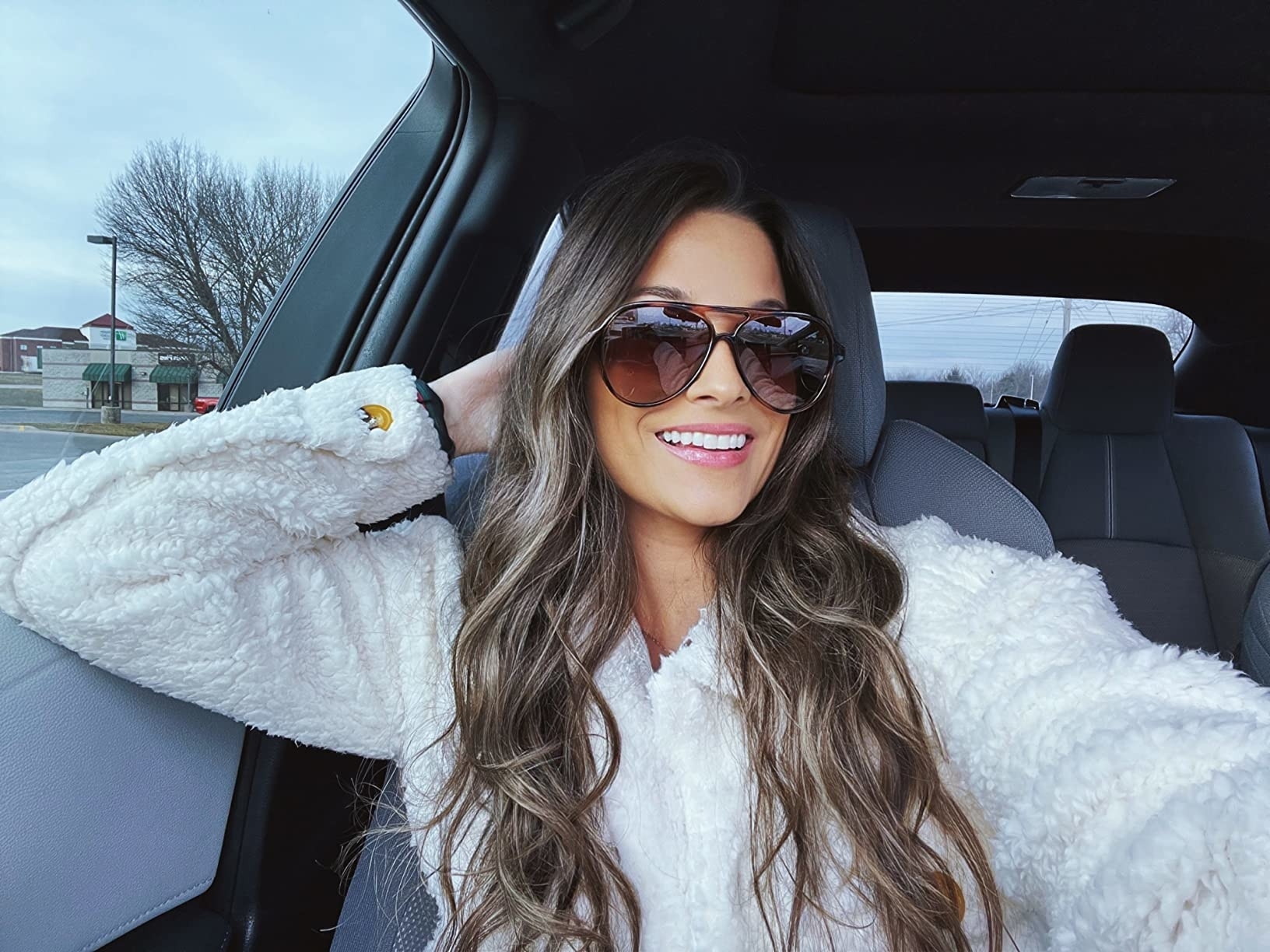 Woman in car wearing sunglasses and a white fluffy jacket smiling at the camera