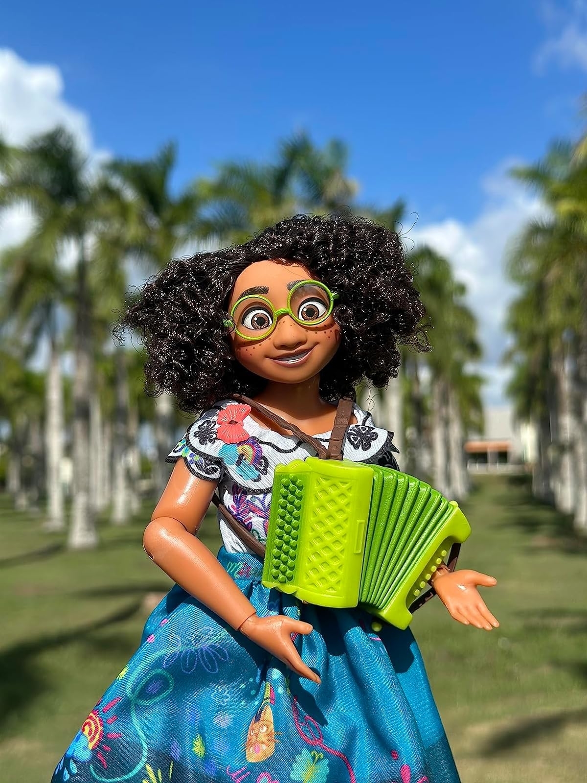Doll character with an accordion, wearing a floral pattern dress and glasses, outdoors with palm trees in the background