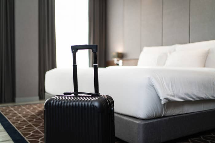 A suitcase stands in front of a neatly made bed in a hotel room, likely suggesting travel or a stay