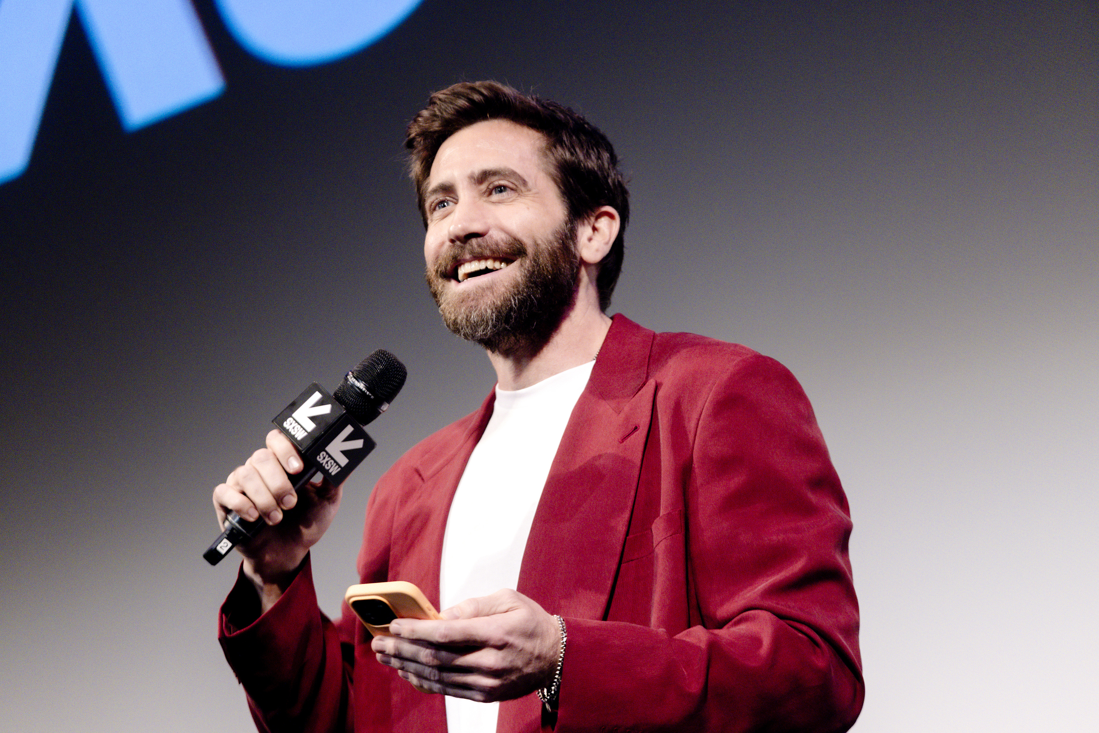 Closeup of Jake Gyllenhaal speaking onstage while holding a smartphone
