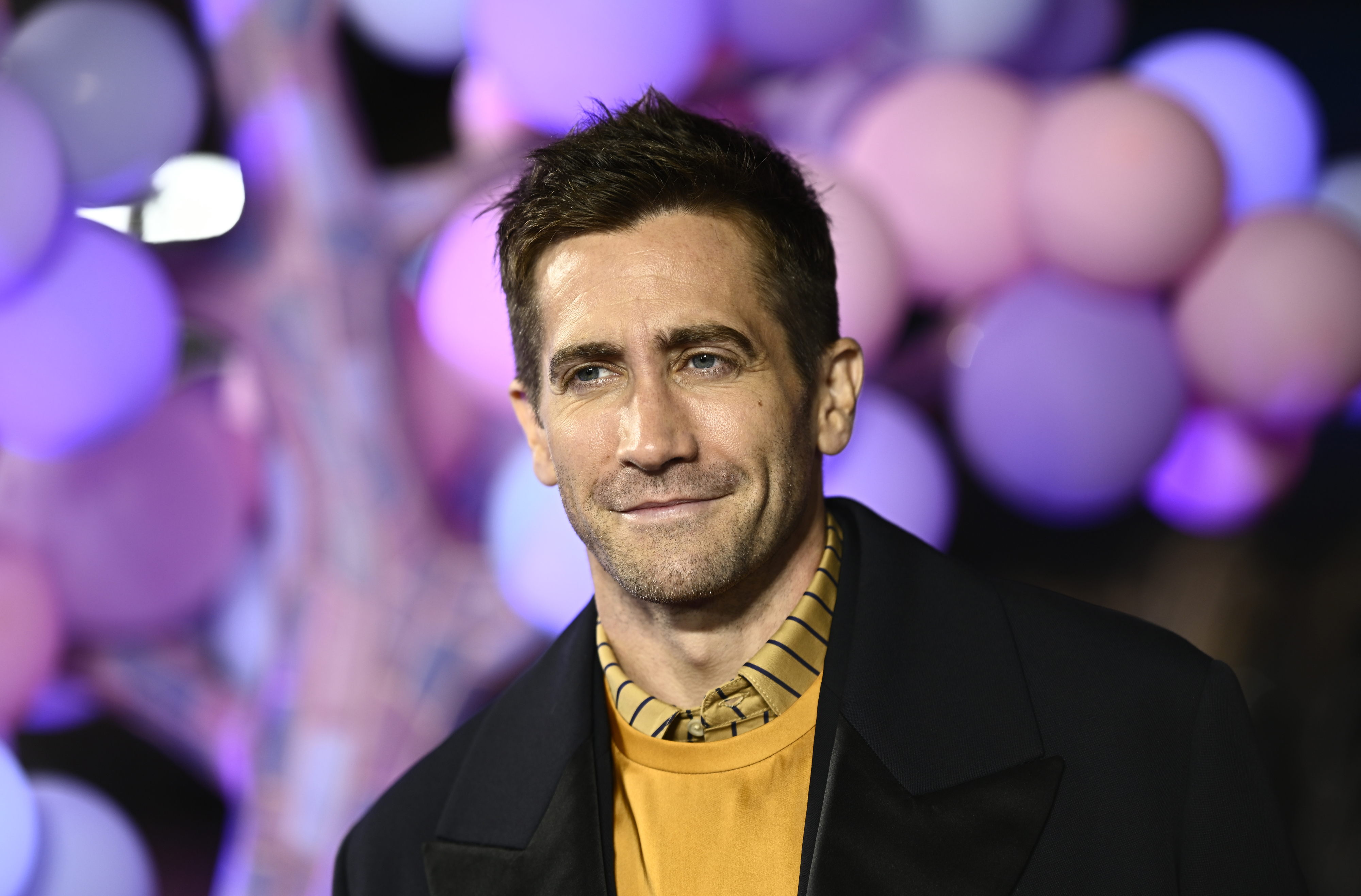 Jake Gyllenhaal in a shirt and jacket, smiling at a premiere with balloons in the background