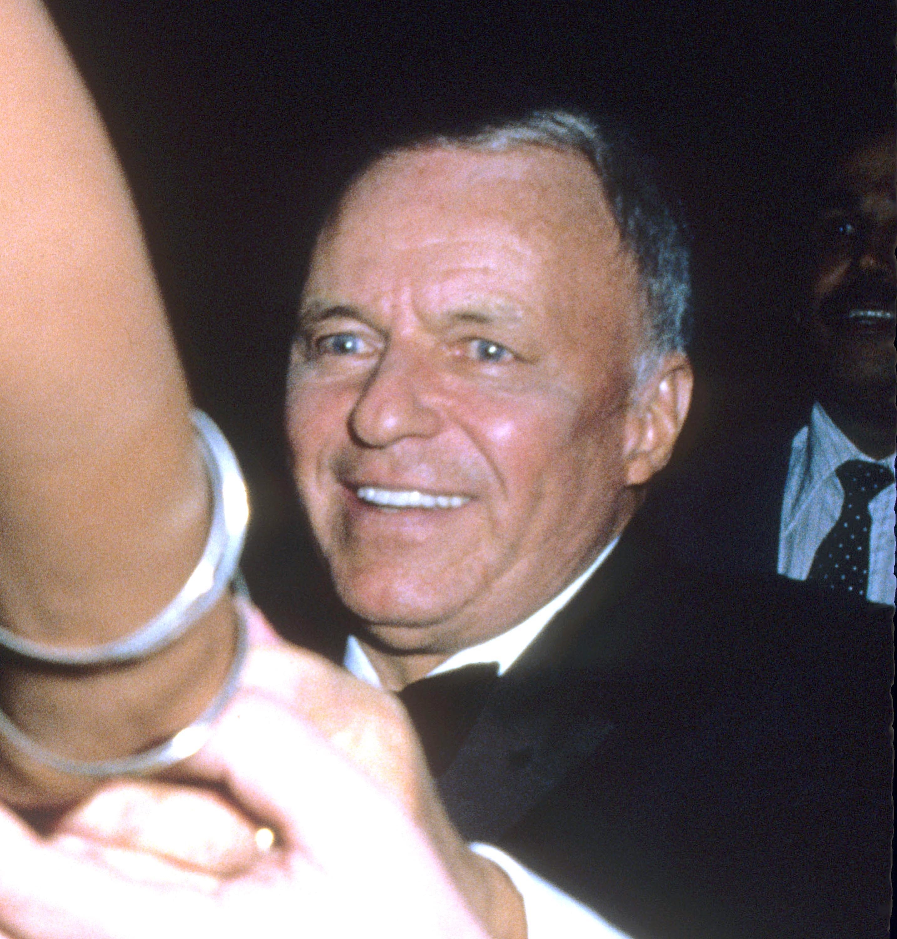Frank Sinatra smiling, wearing a tuxedo in a crowded event