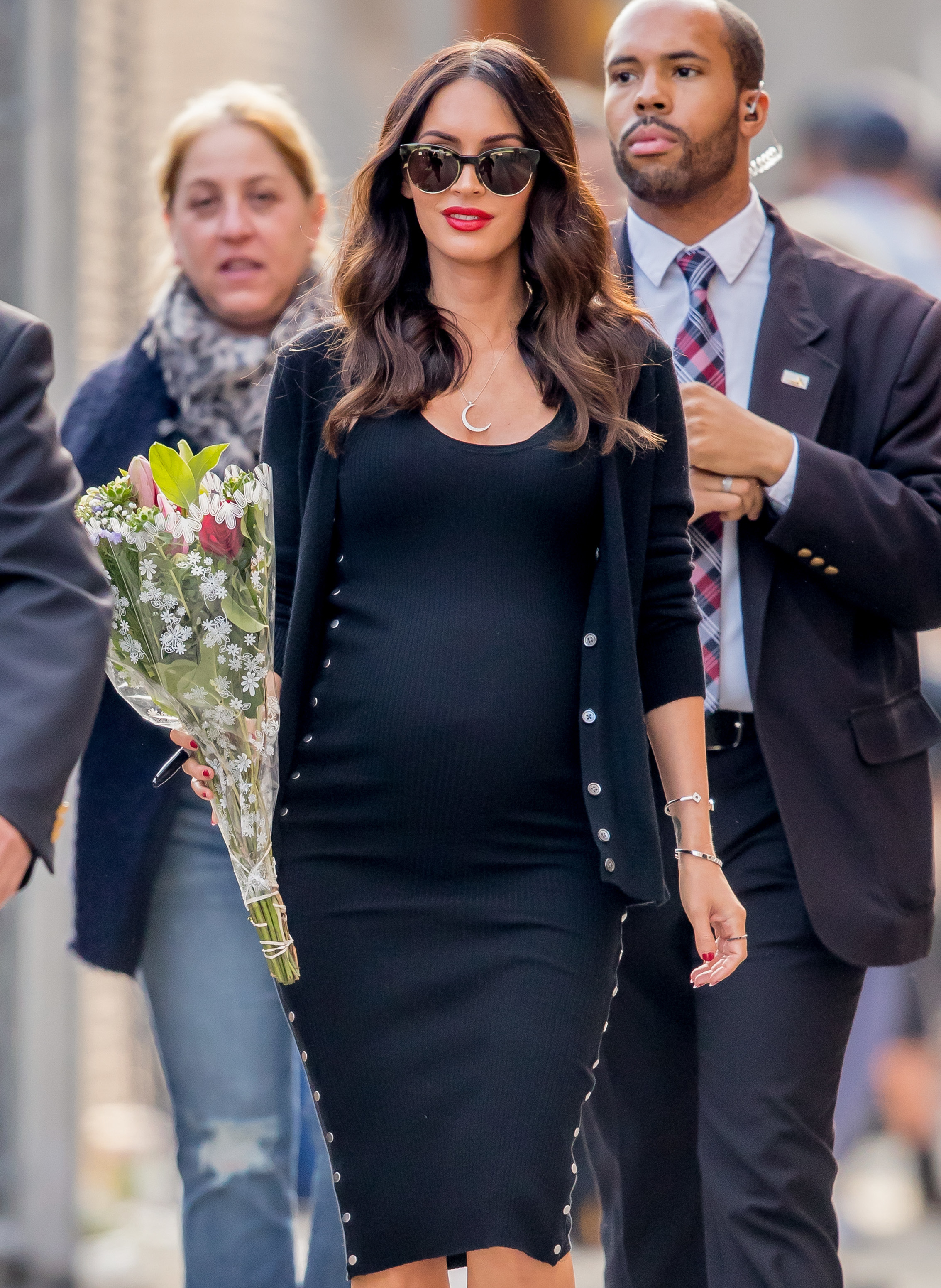 Pregnant Megan Fox in a snug black dress with a cardigan, holding flowers, flanked by security personnel