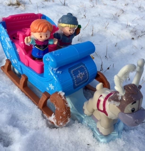 Two toy figures riding a sleigh with a reindeer toy in the snow, evoking winter playtime