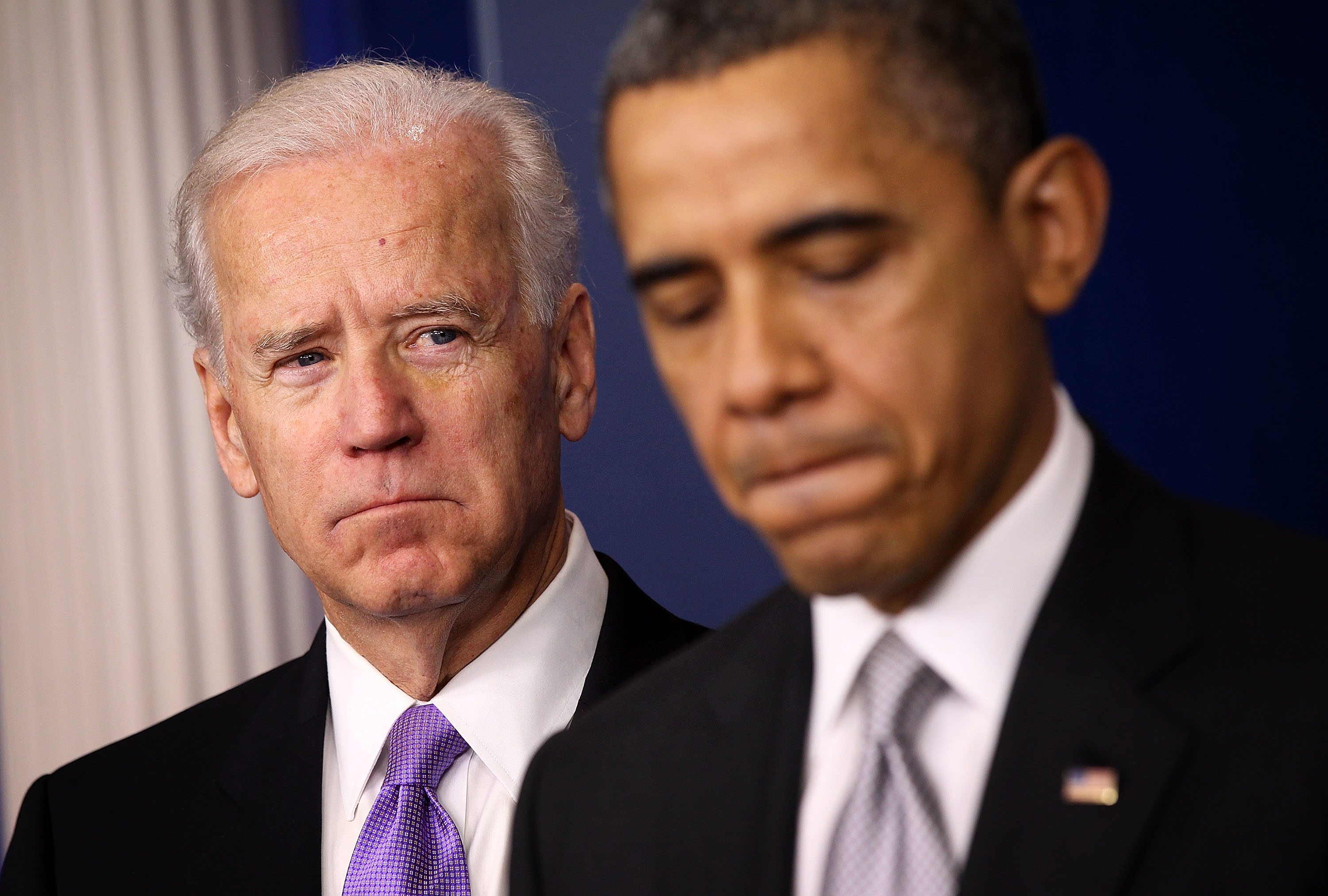 Joe Biden and Barack Obama standing, both in business suits with serious expressions