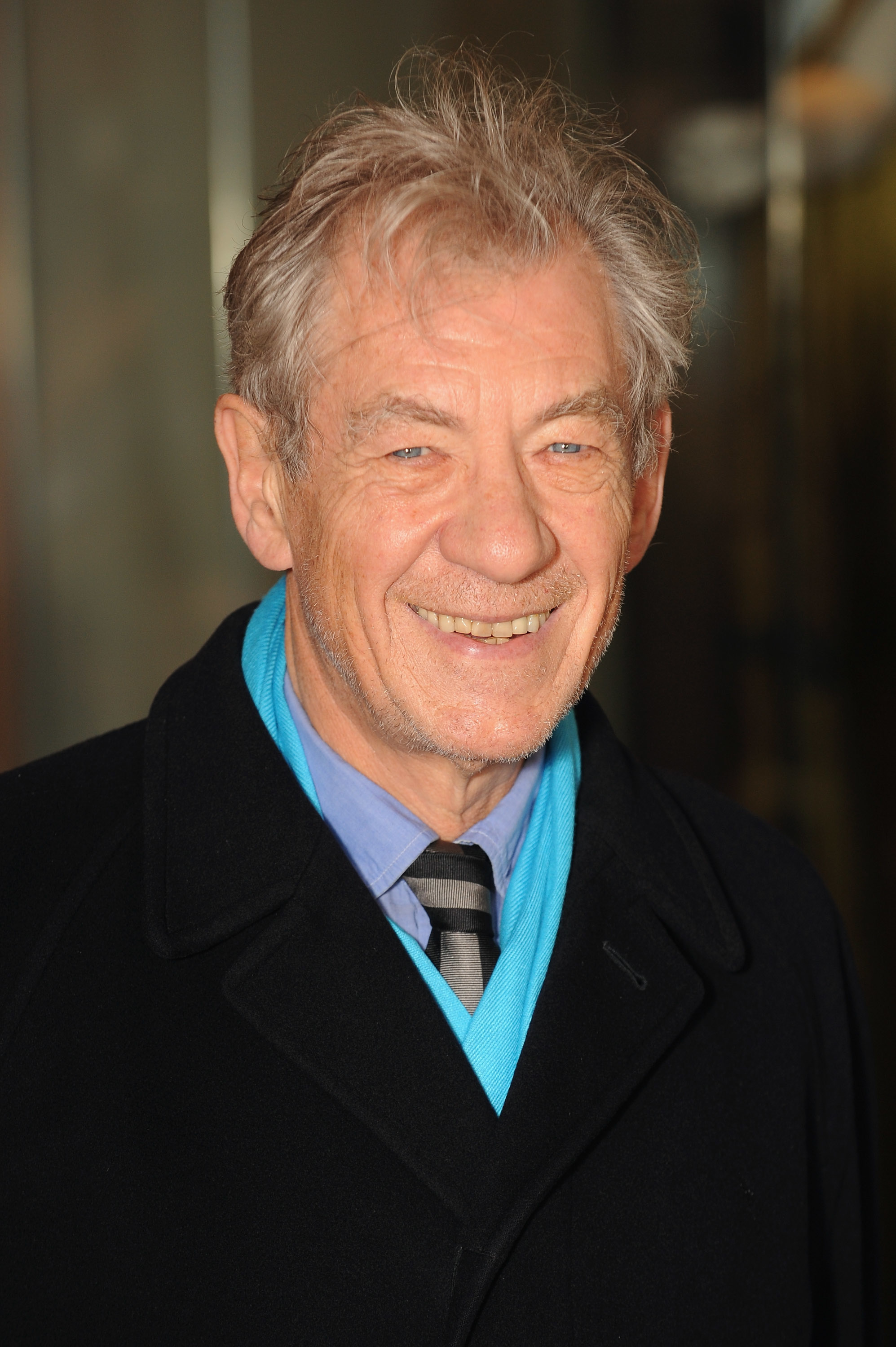 Sir Ian McKellen in a black coat and blue scarf, smiling at an event