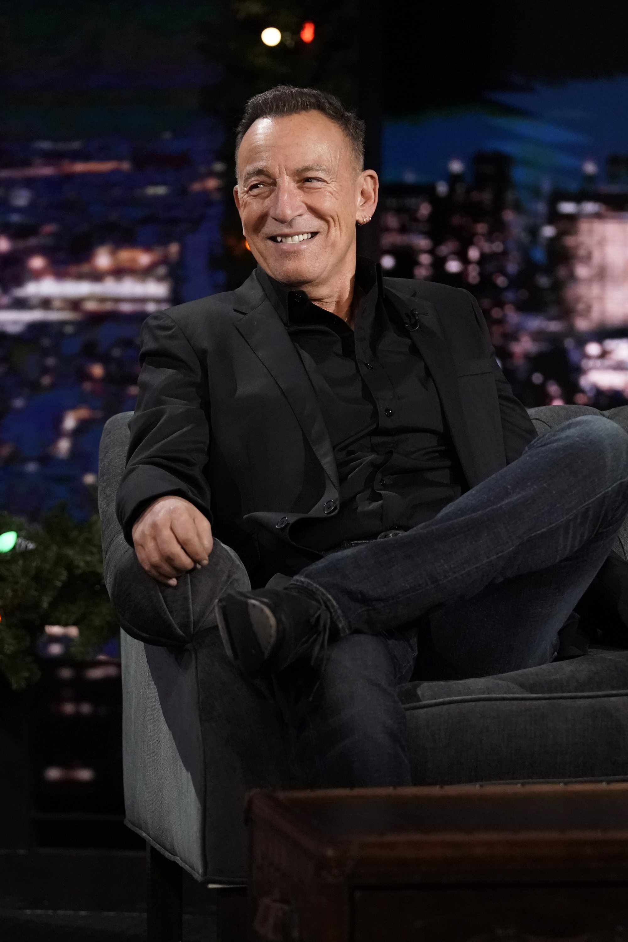 Celebrity male sitting cross-legged in a talk show setting wearing a black shirt and jacket