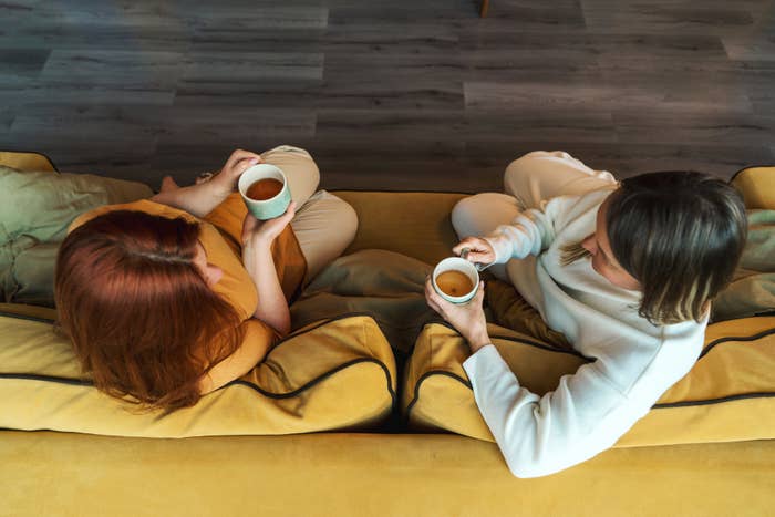 Two individuals are sitting on a couch, viewed from above, each holding a cup, engaged in a conversation