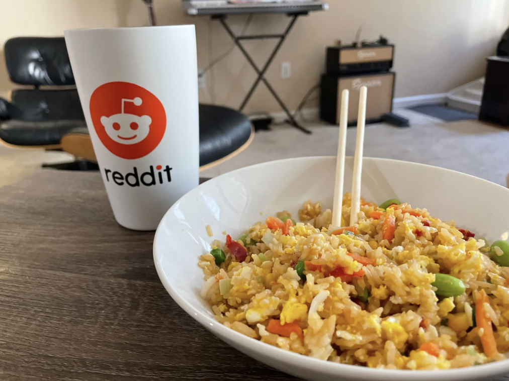 A bowl of fried rice with vegetables next to a Reddit-branded cup on a table