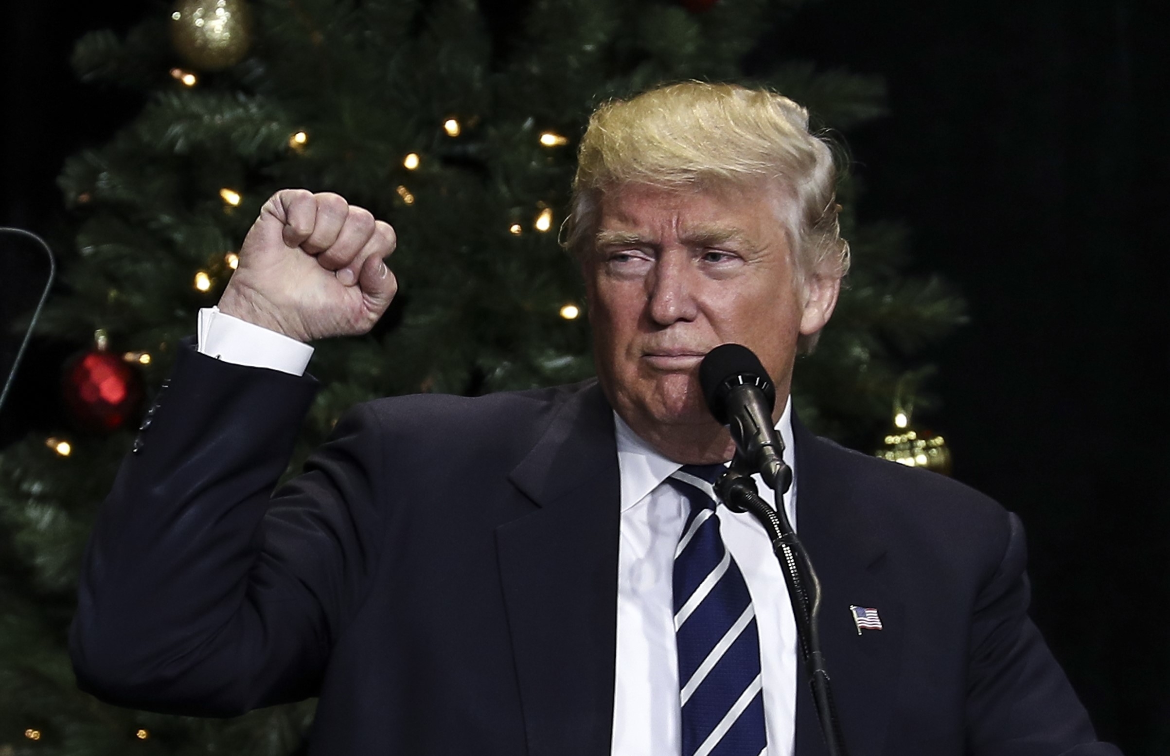 Donald Trump, wearing a suit, makes a gesture at a podium with a Christmas tree in the background
