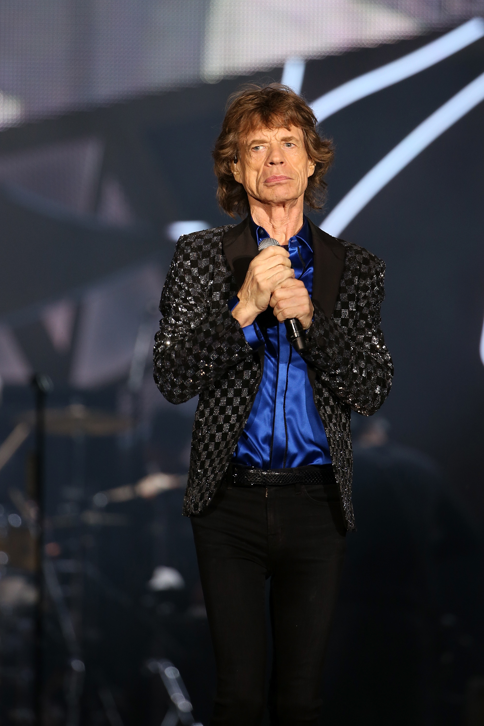 Mick Jagger onstage in a sparkling black jacket, blue shirt, holding a microphone