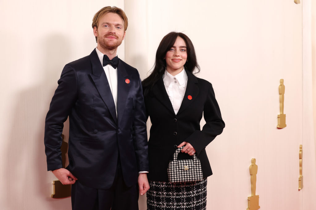 Billie and Finneas standing side by side: one in a black suit with a lapel pin, the other in a black blazer, white shirt, and carrying a checkered bag