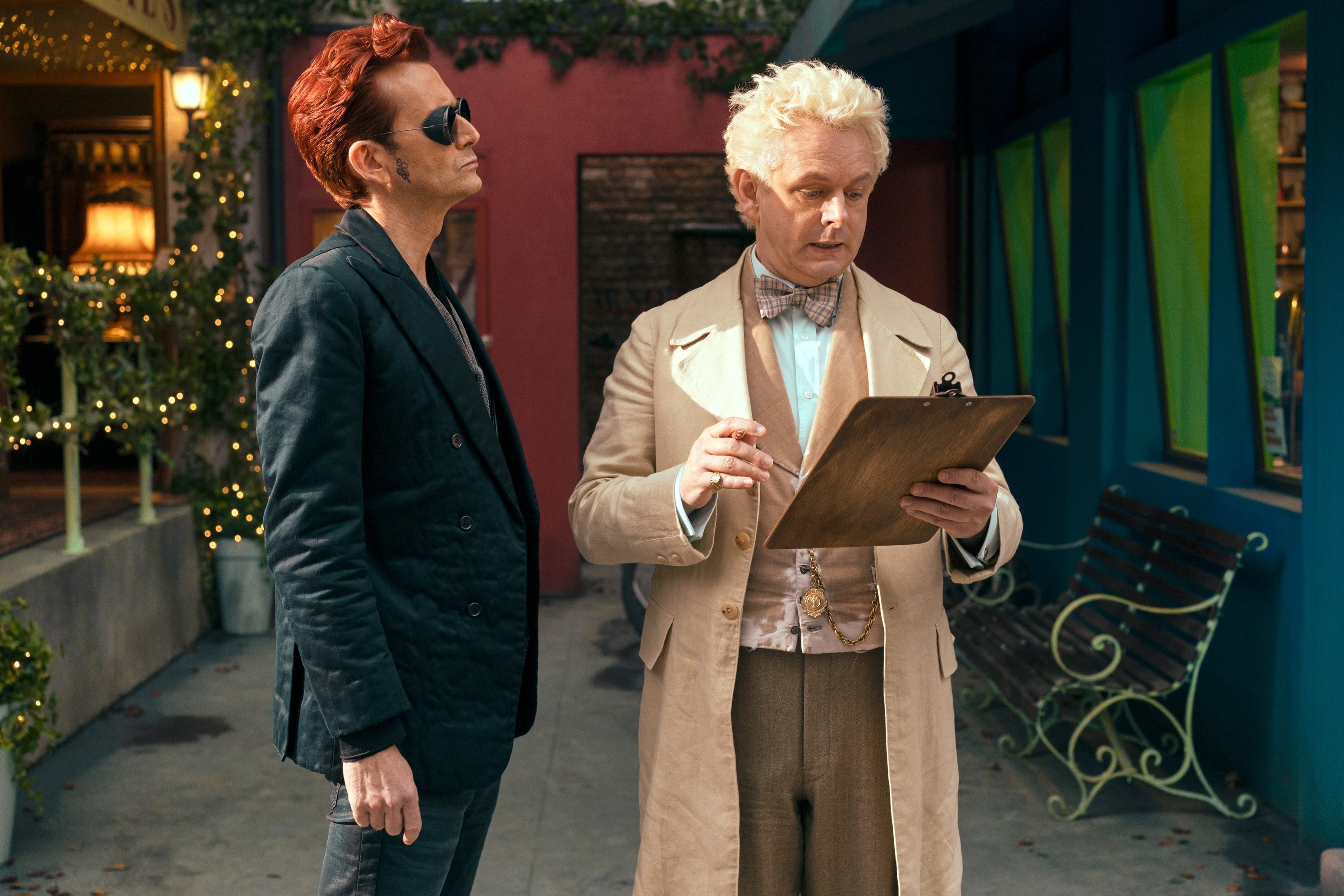 Two characters from &quot;Good Omens,&quot; Crowley in aj acket and Aziraphale in a bow tie, examine a clipboard