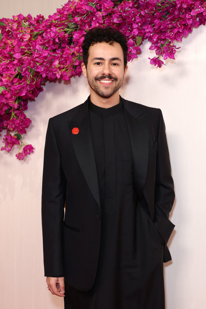 Man smiling, wearing a sleek black suit with a red poppy pin, standing before a floral backdrop