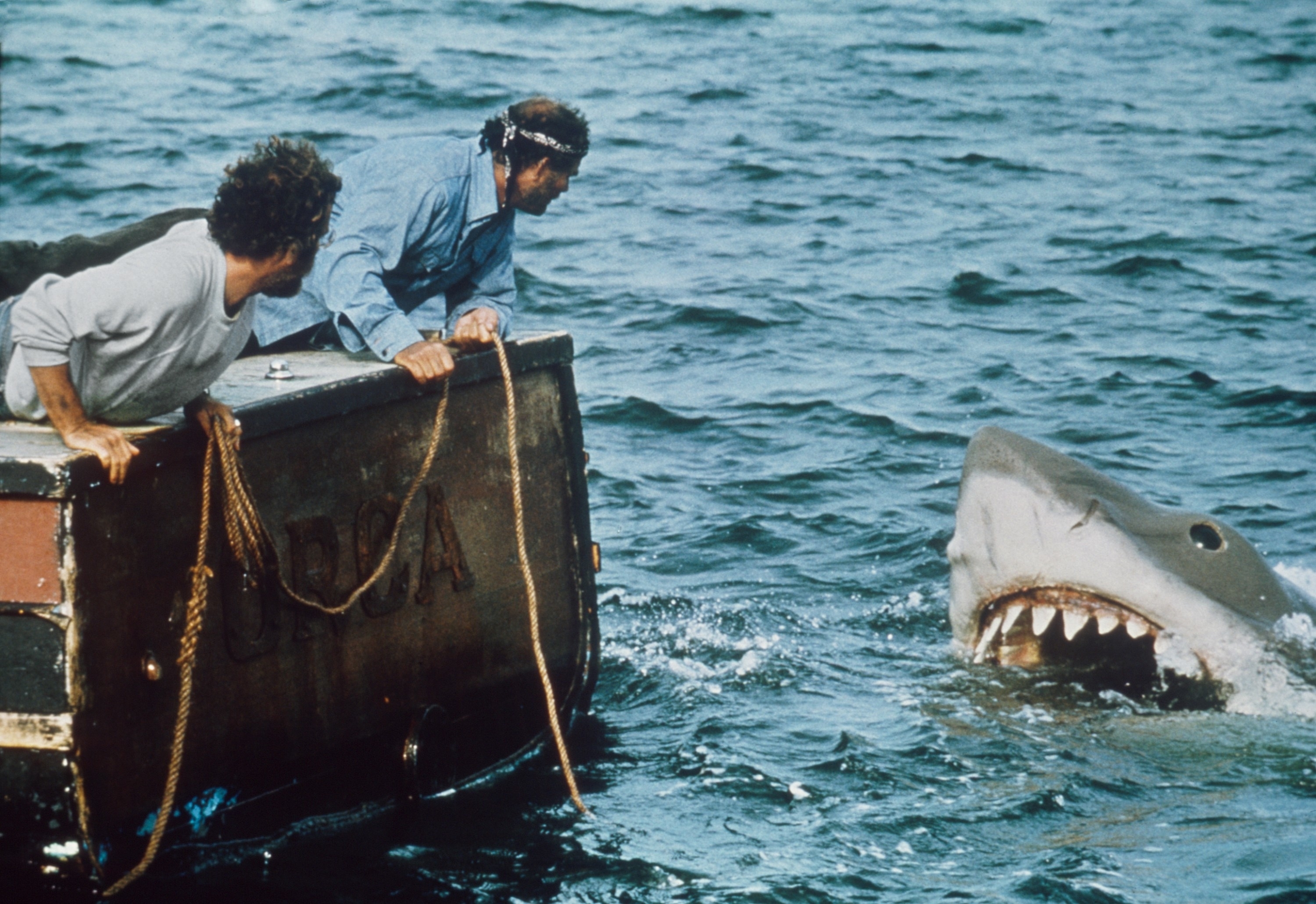 Two characters from the movie Jaws are on a boat, looking apprehensively at a large mechanical shark emerging from the water