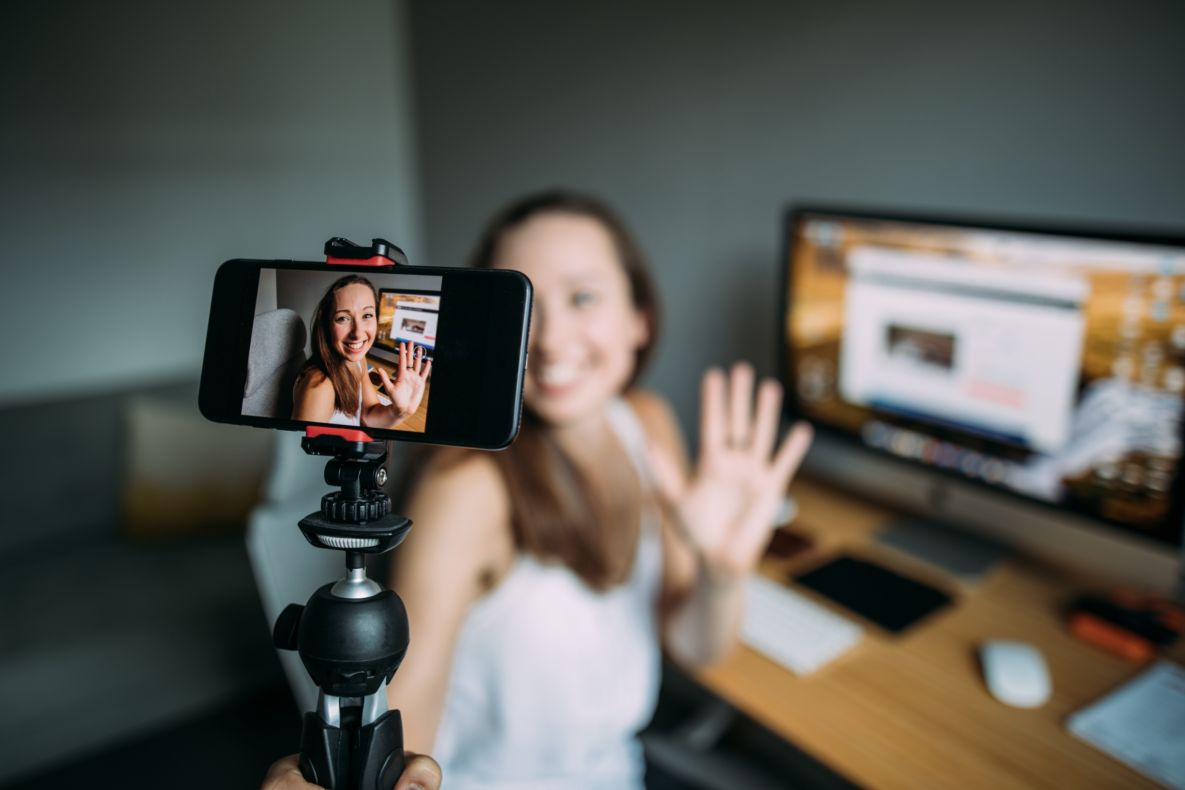 Woman recording a video at a desk with a smartphone mounted on a tripod. She&#x27;s smiling and waving at the camera