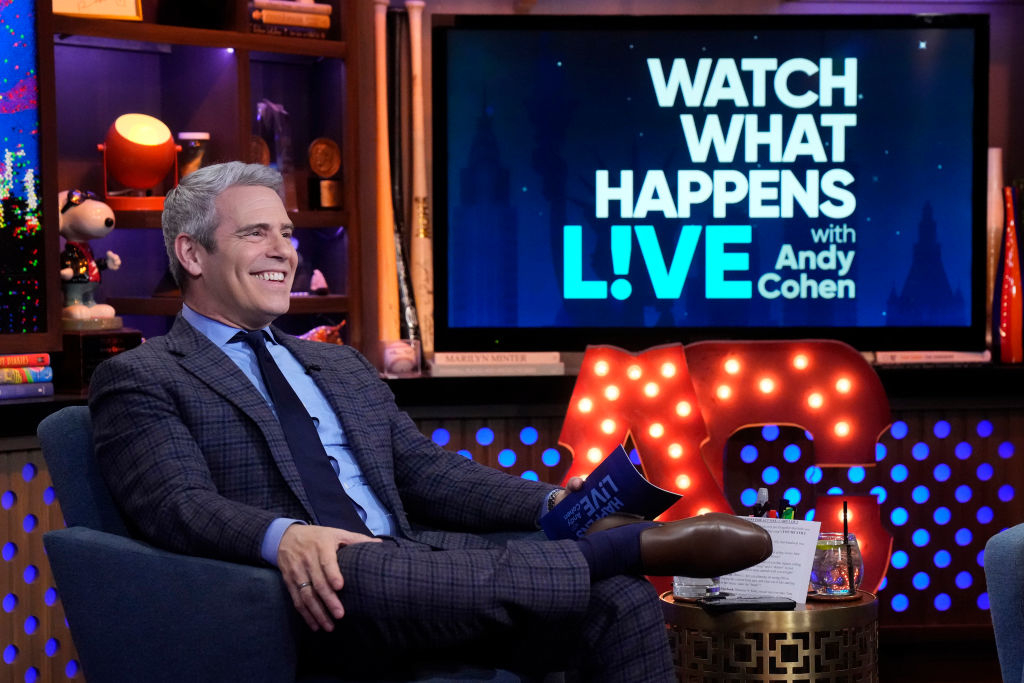 Andy Cohen is seated smiling on the set of &quot;Watch What Happens Live,&quot; holding cue cards