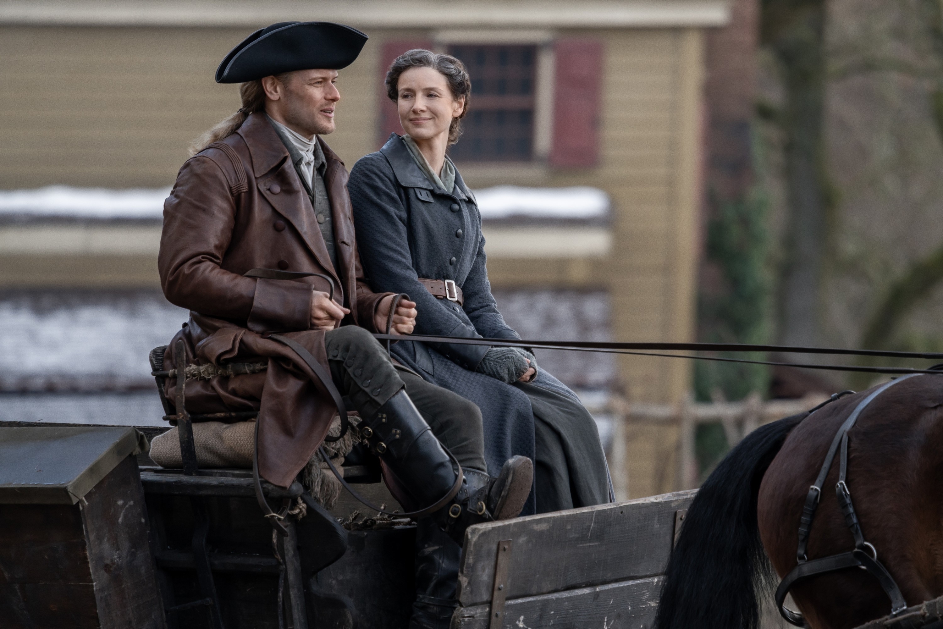 Jamie and Claire are sitting in a horse-drawn carriage, dressed in period costumes