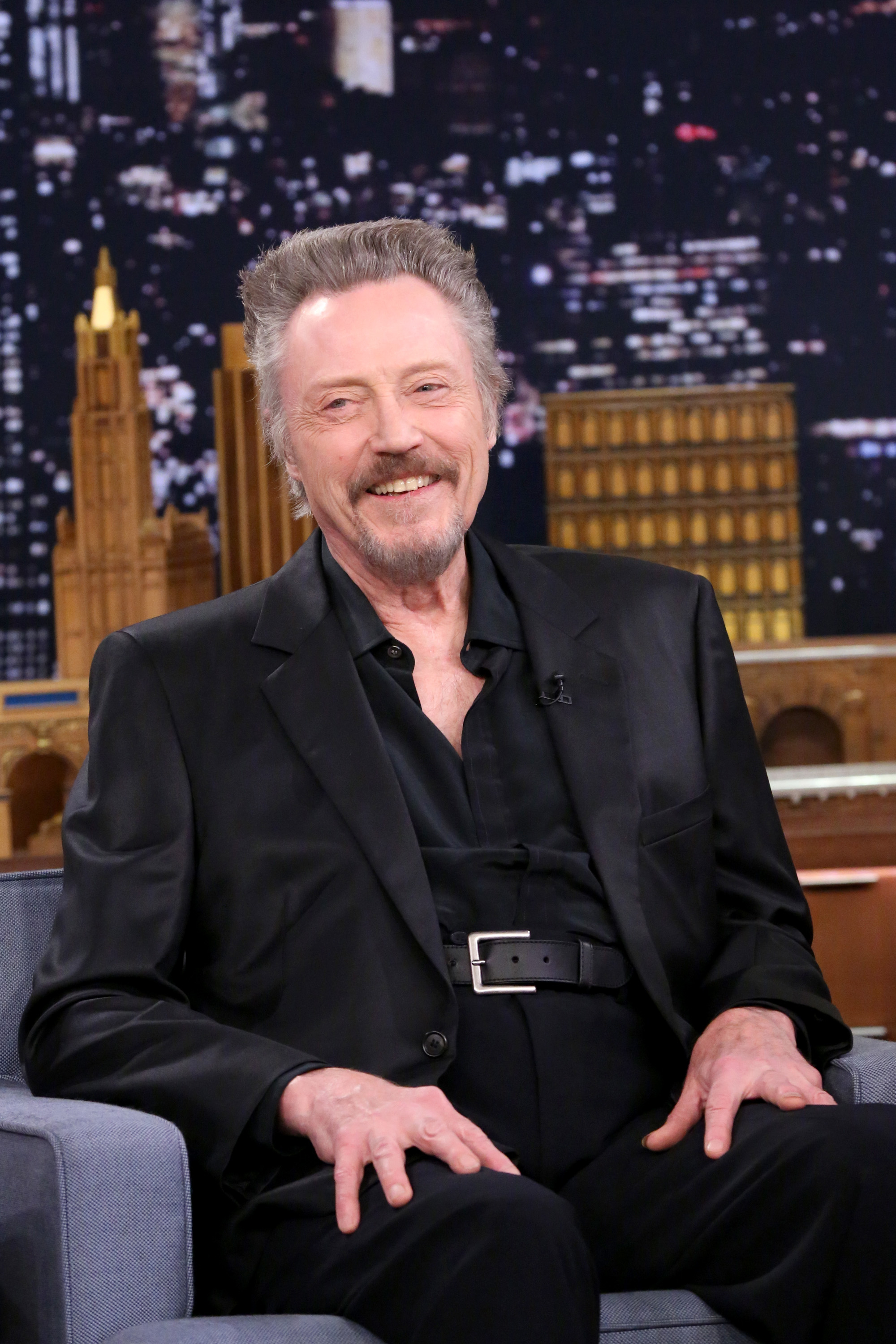 Man in black suit sitting, smiling on talk show set