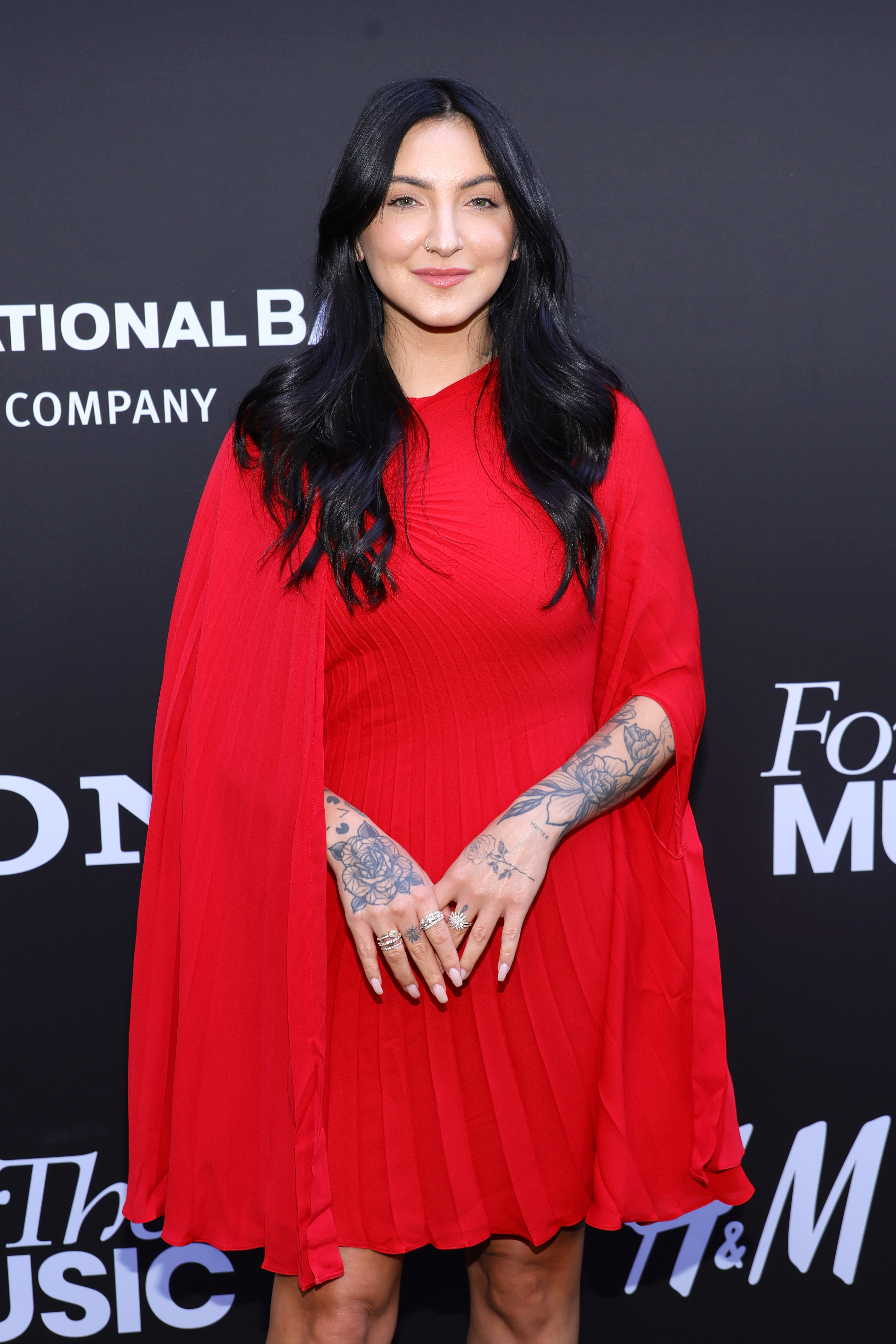 Julia in a red cape-style dress posing with hands together, showing sleeve tattoos