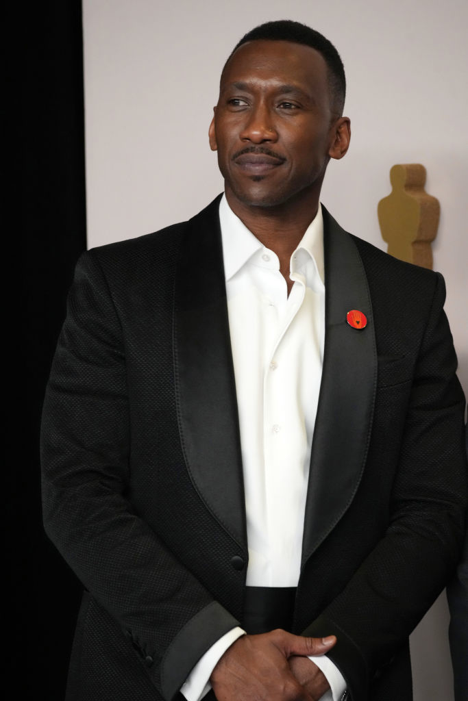 Mahershala Ali wearing a tailored suit with a white shirt and a red accessory on the lapel