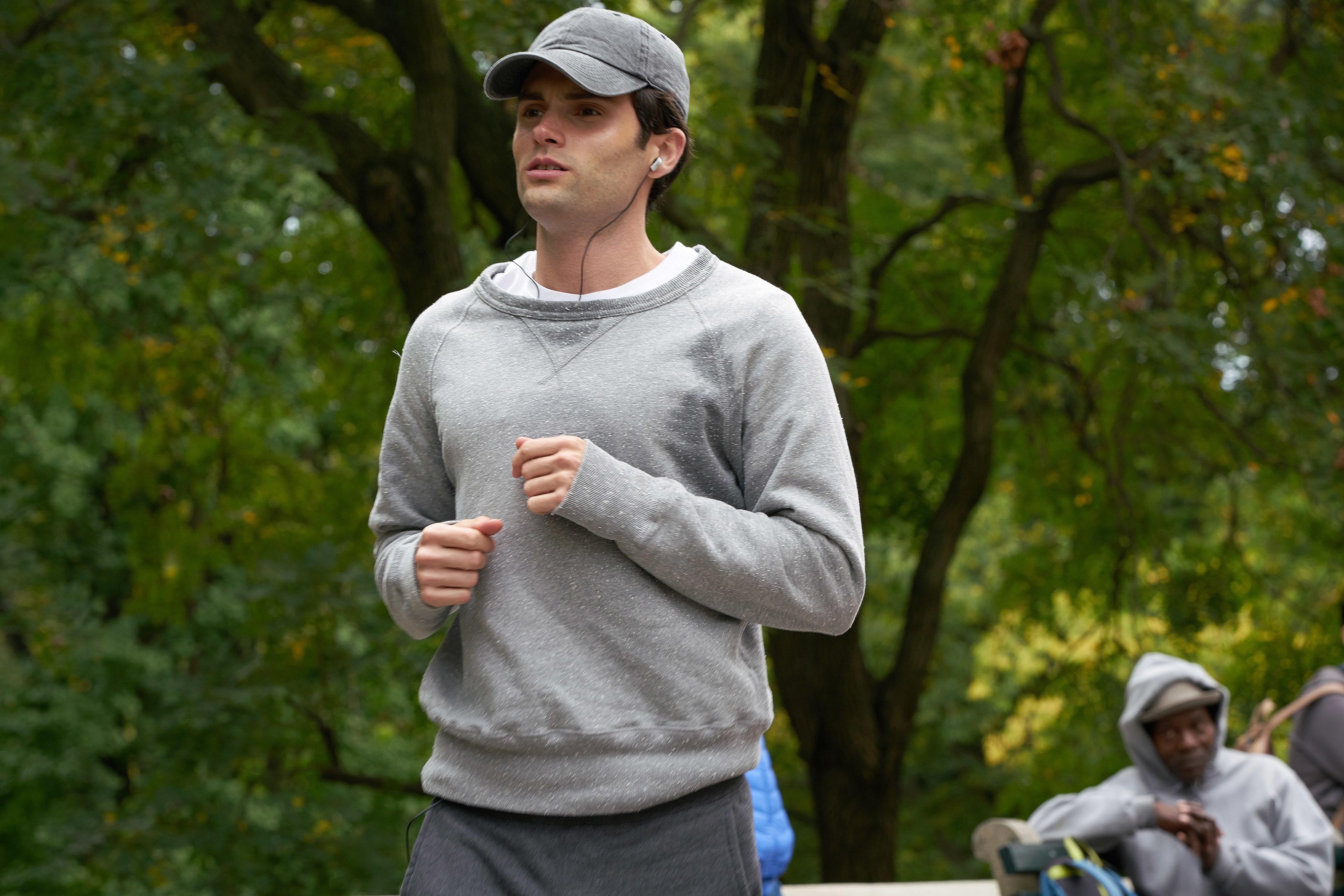Joe in a hoodie and baseball cap on a jog through a park with onlookers