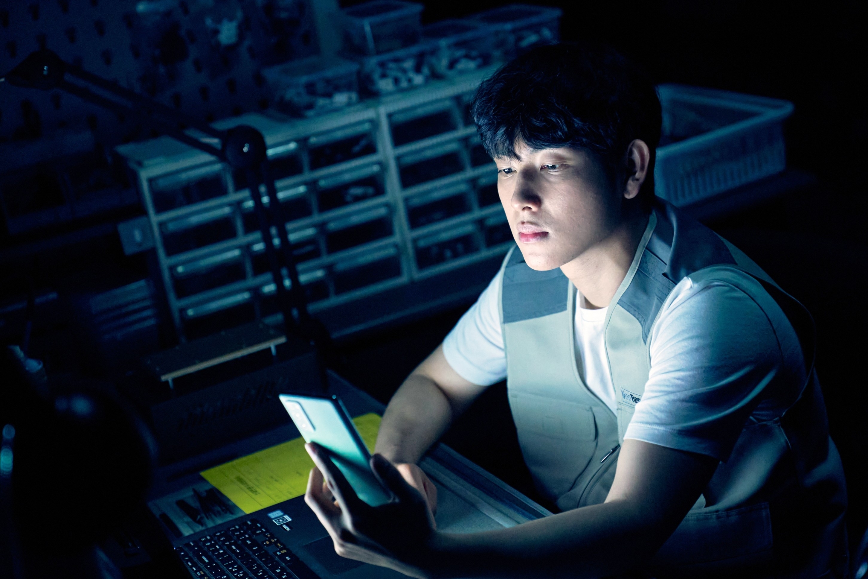 Oh Jun-yeong in a dark room looking at his phone screen with focus, laptop open nearby