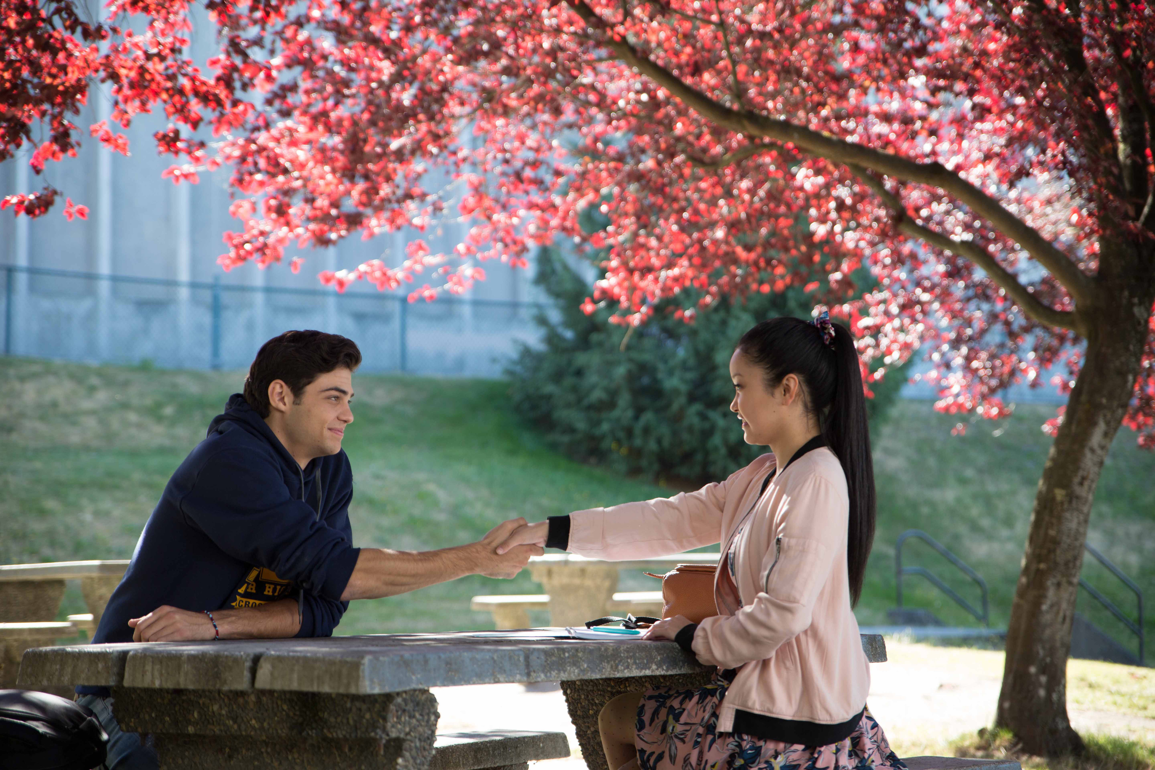 Peter and Lara Jean, sitting at a stone table under a flowering tree, reaching out for a handshake