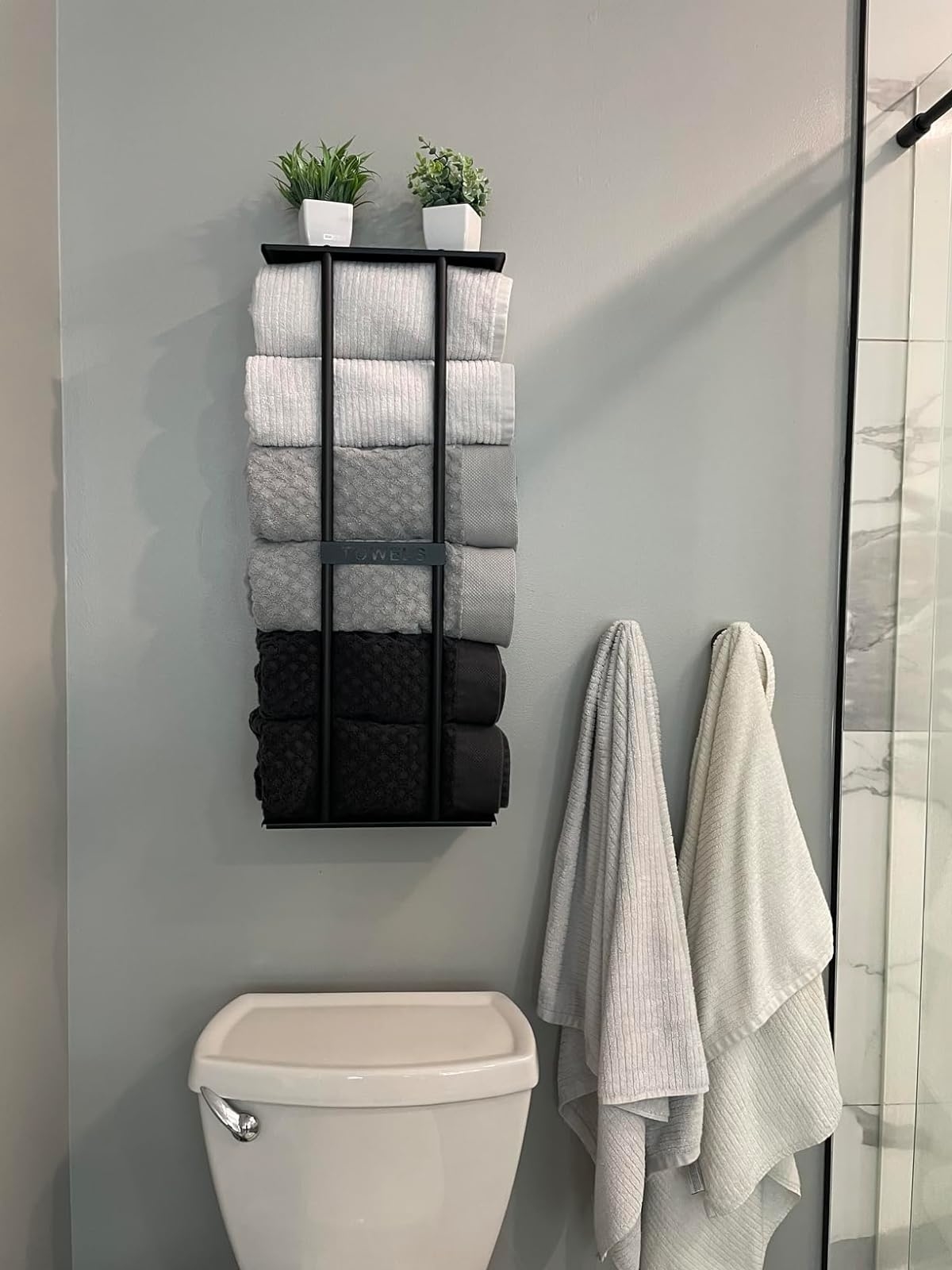 A wall-mounted towel rack with variously sized gray and black towels, above a toilet, with two small plants on top