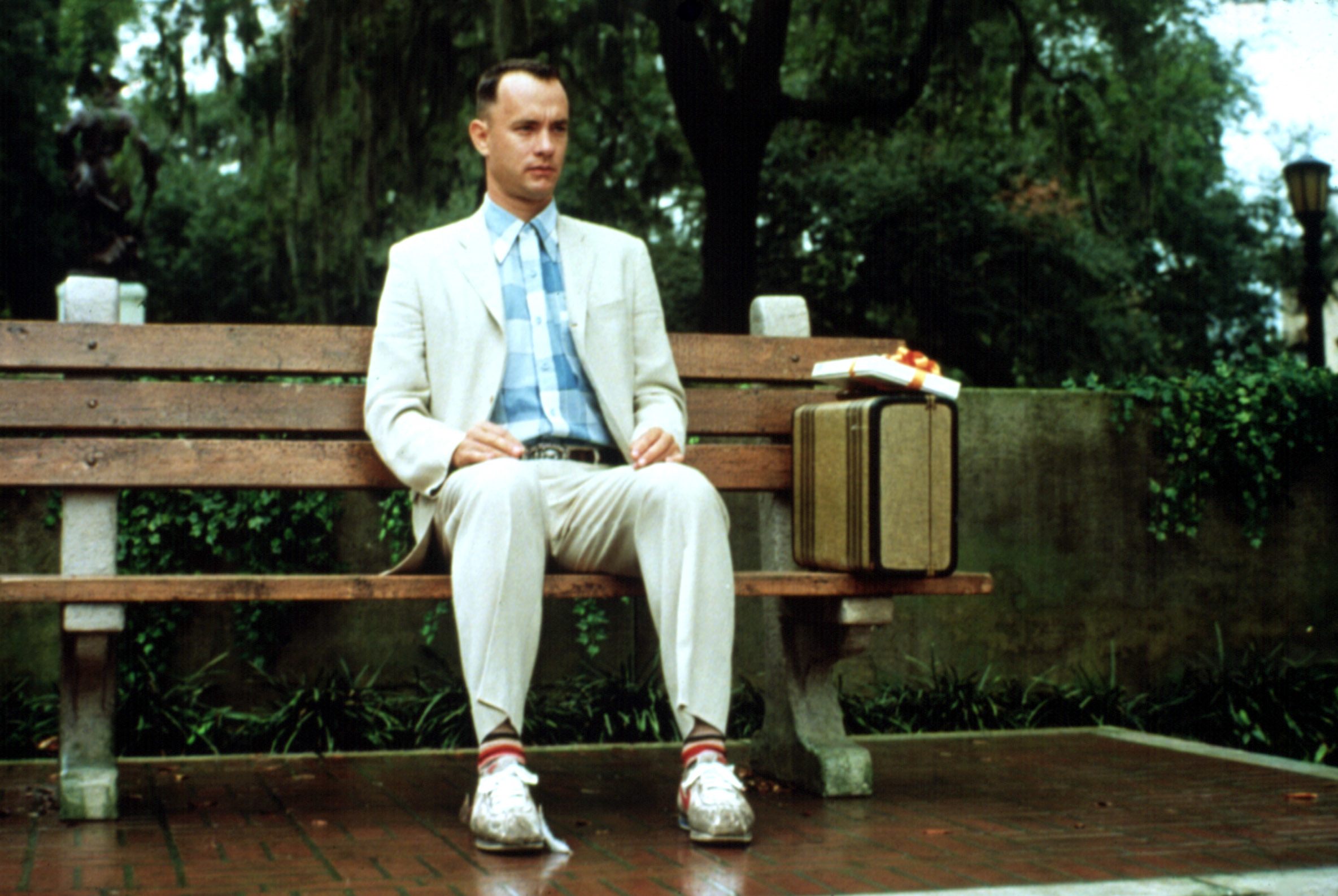 Tom Hanks as Forrest Gump, seated on a bench with a suitcase, wearing a suit and sneakers