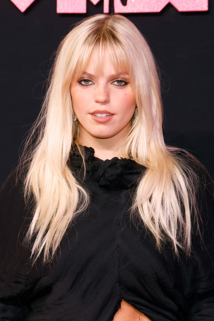 Person poses on red carpet in a black ruffled outfit, with long blonde hair and bangs