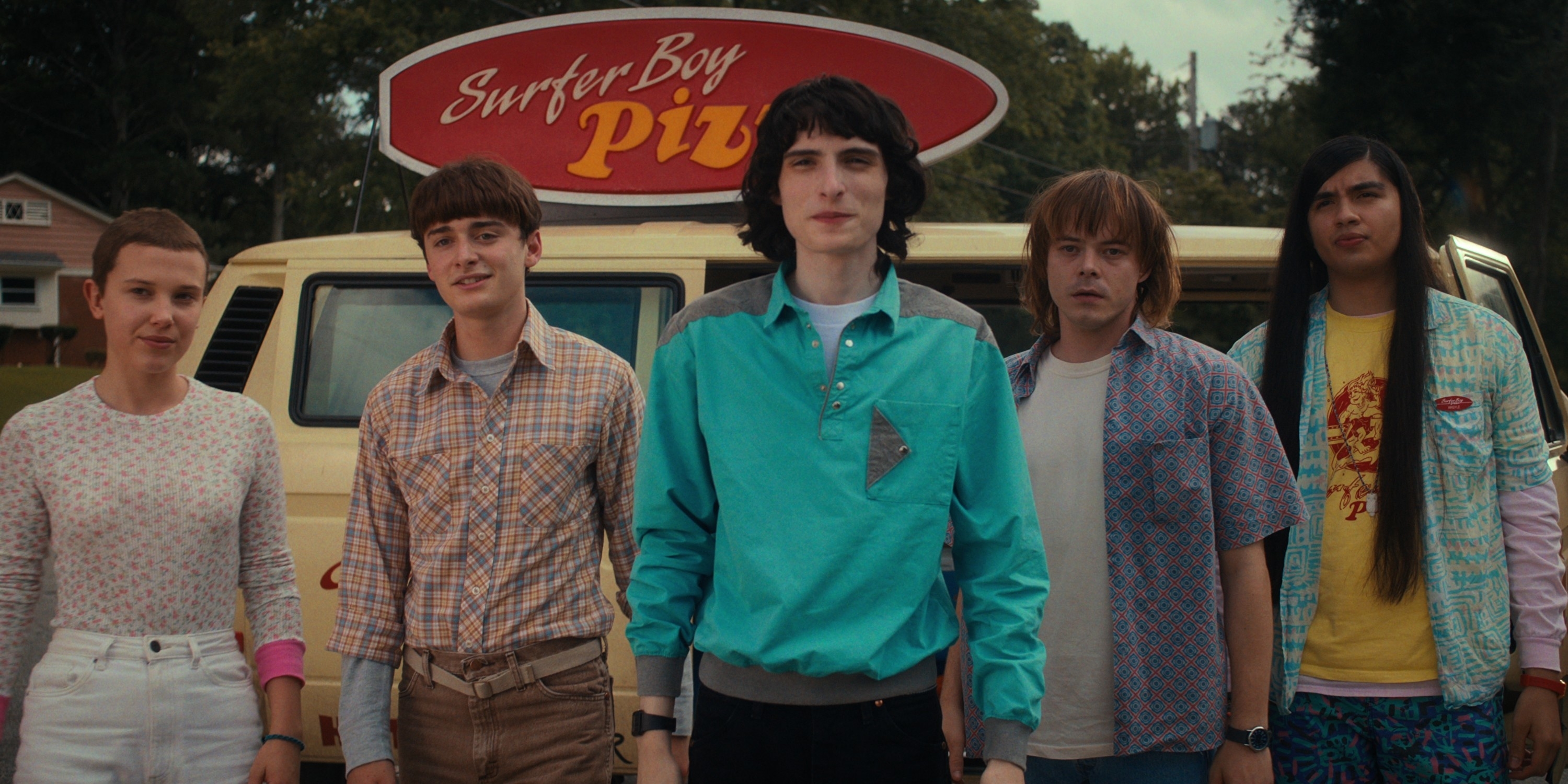 Characters from the show &#x27;Stranger Things&#x27; standing in front of Surfer Boy Pizza. They are dressed in 80s casual wear