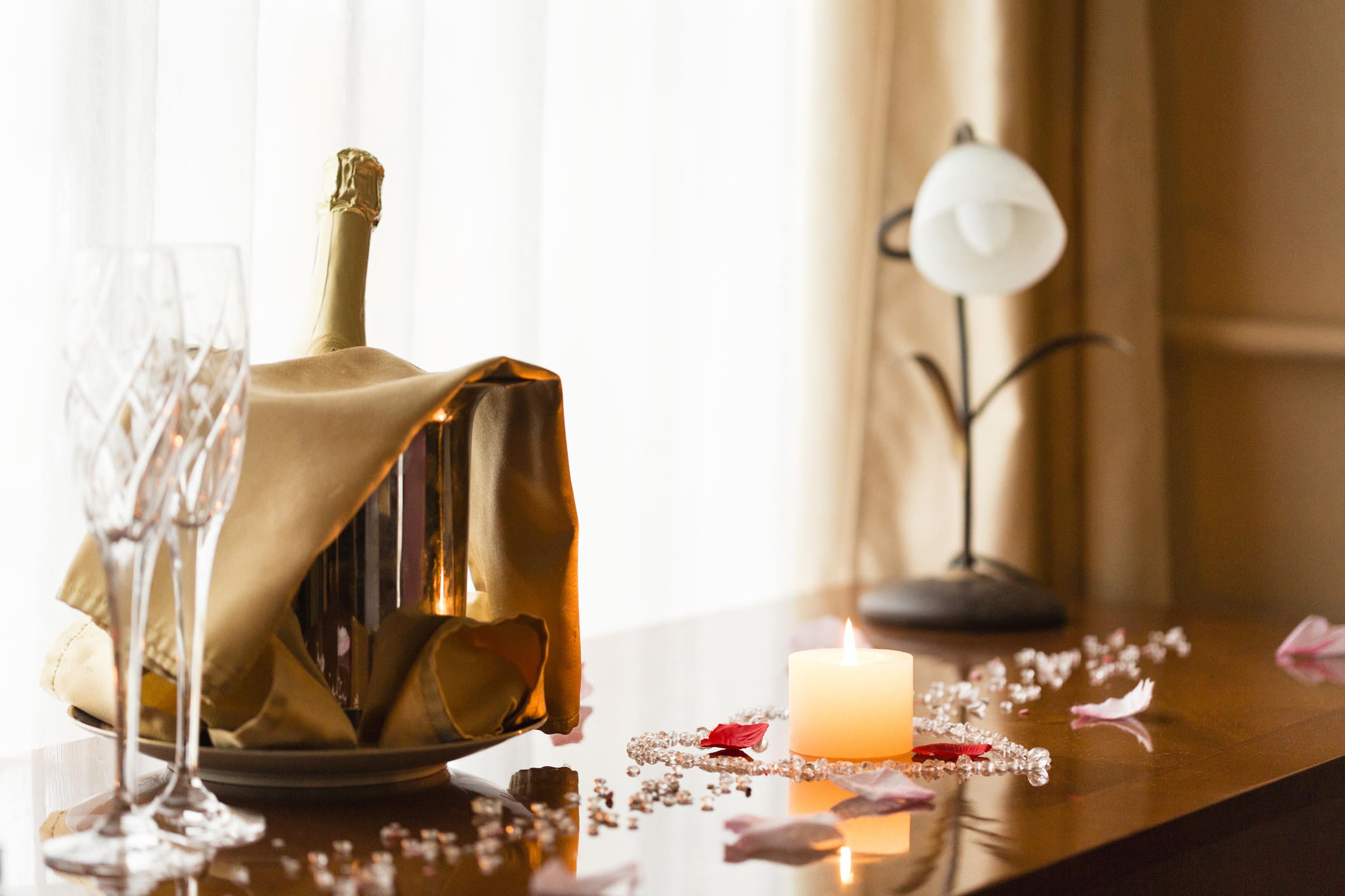 Champagne bottle in gold wrapper on table with two glasses, candle, and rose petals for a wedding setting