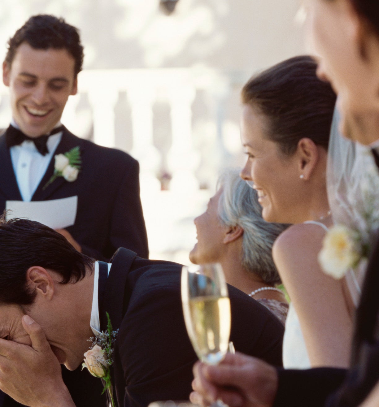 Groom laughing with hand on face, bride smiling beside him, man giving speech at wedding