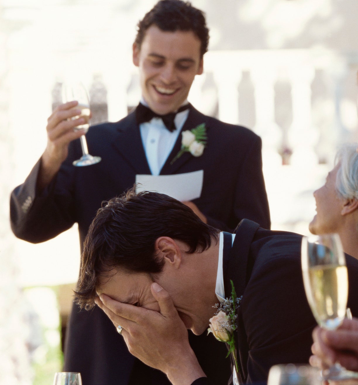 Groom laughing with hand on face, bride smiling beside him, man giving speech at wedding
