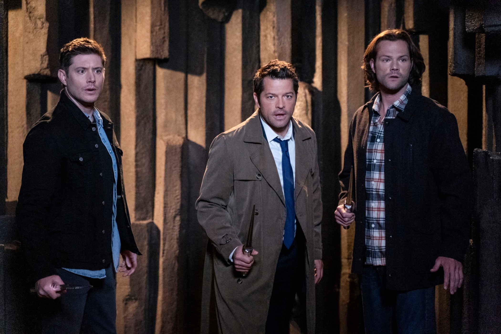 Dean, Castiel, and Sam from &quot;Supernatural&quot; in character, standing alert in a dimly lit wooden room