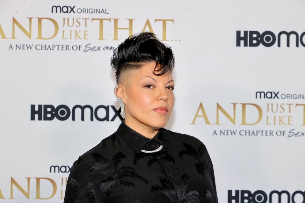 Person posing at an event with an HBO Max backdrop, wearing a black embossed outfit with a high collar