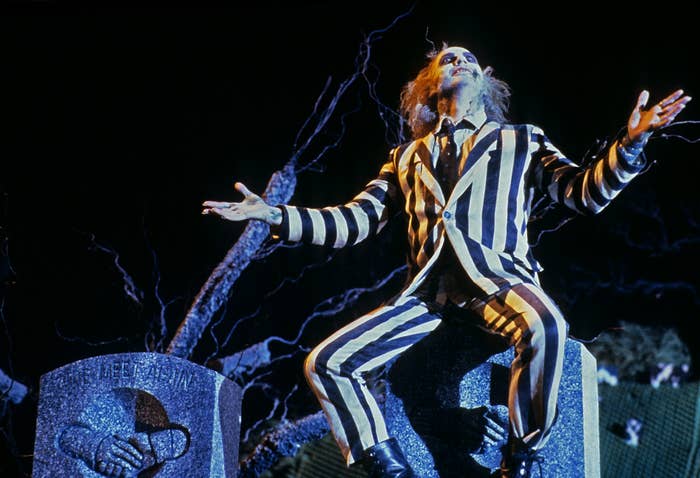 Beetlejuice character in striped suit stands with arms outstretched as he sits on a tombstone
