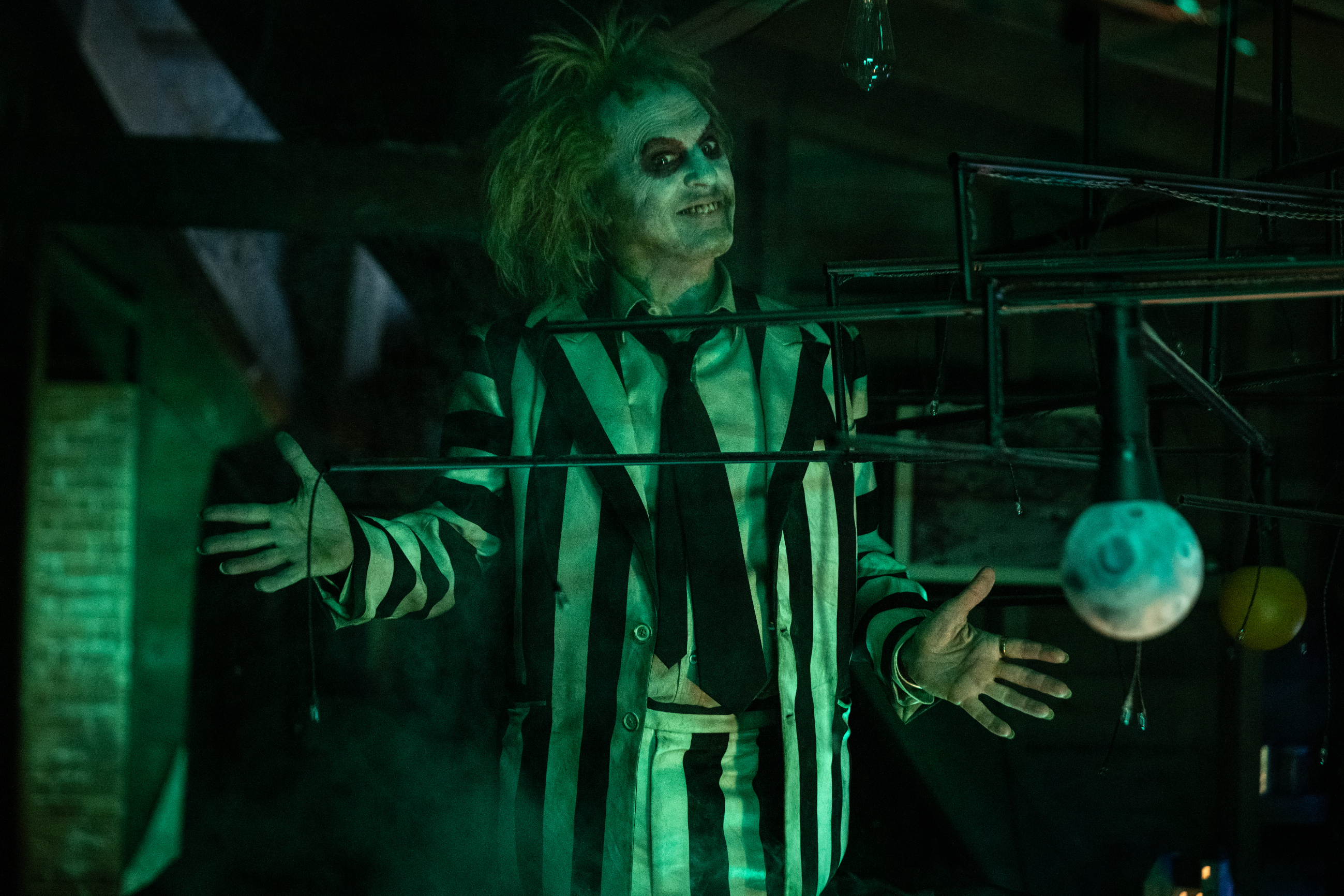 Beetlejuice in his signature striped suit with wild hair, outstretched arms, and dramatic makeup, in a dimly lit scene