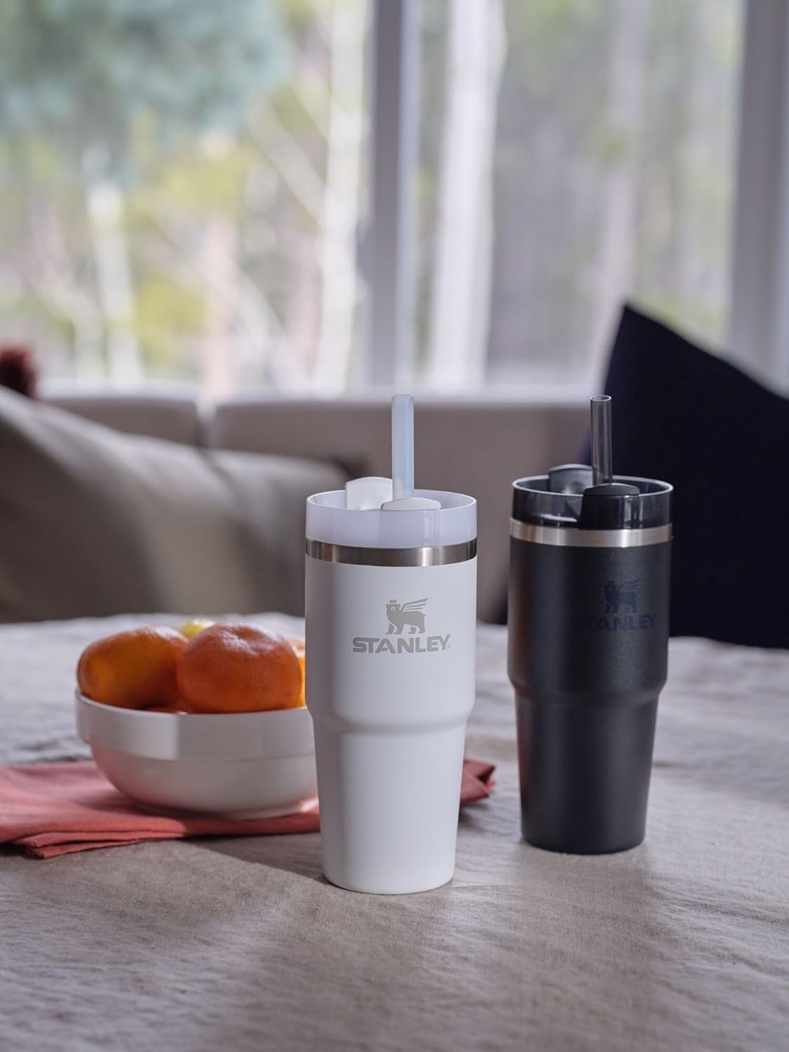 Two Stanley insulated tumblers on a table, one white and one black, with straws. Bowl of oranges in the background