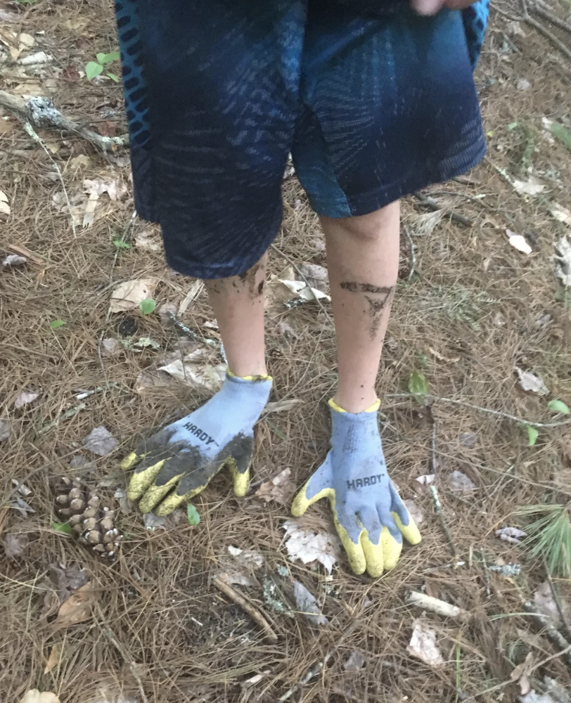 Person standing in a forested area, wearing gloves and shorts with visible dirt on skin and clothing