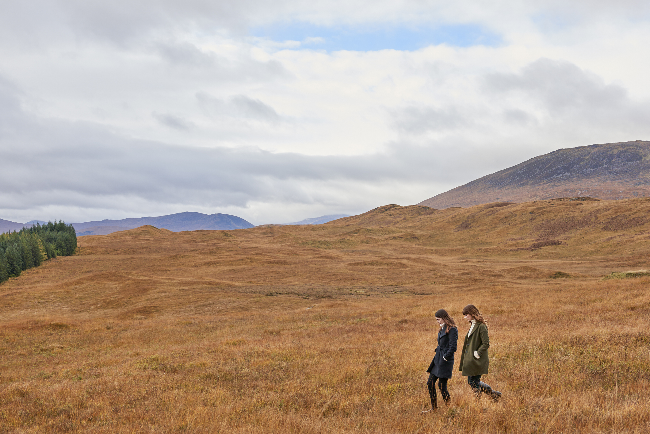 Two people walking through a vast, open field with gentle hills in the background