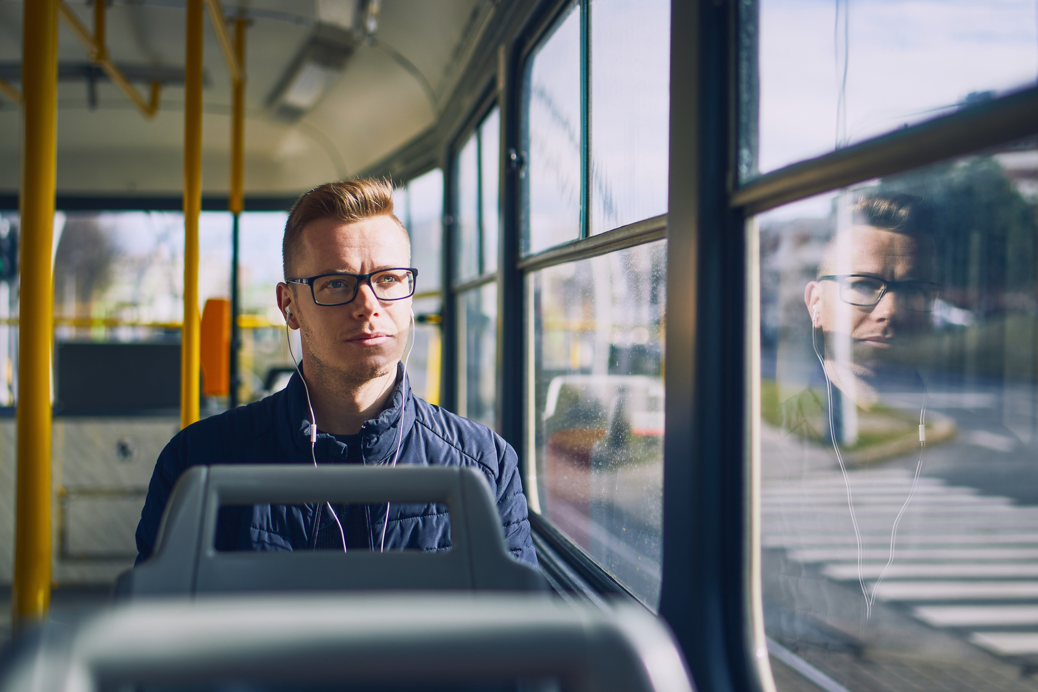 Man wearing glasses sitting inside a bus, looking at the camera, with reflection on the window