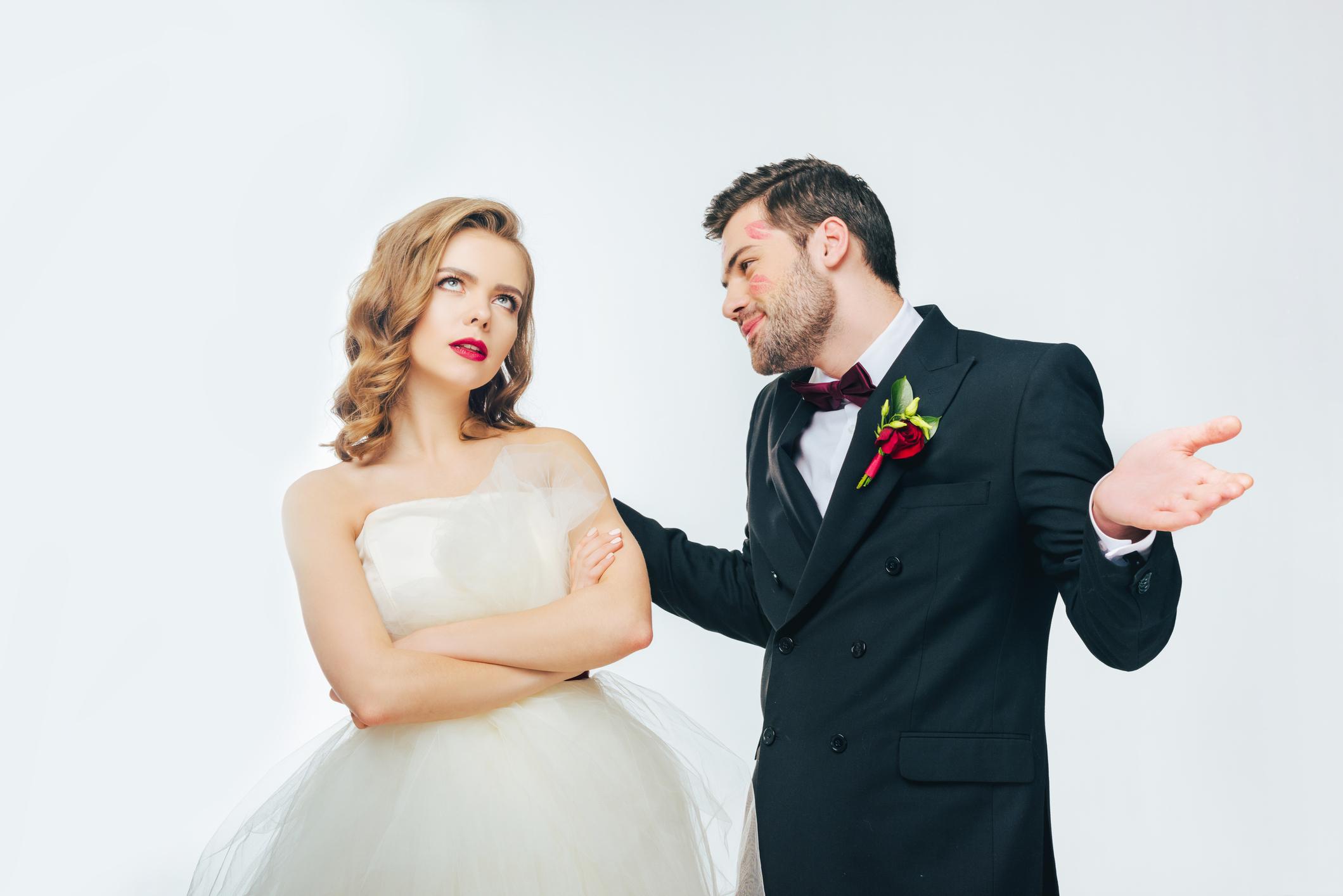 Bride in a strapless gown looks skeptically at groom in a tuxedo with a boutonniere who seems to be explaining something