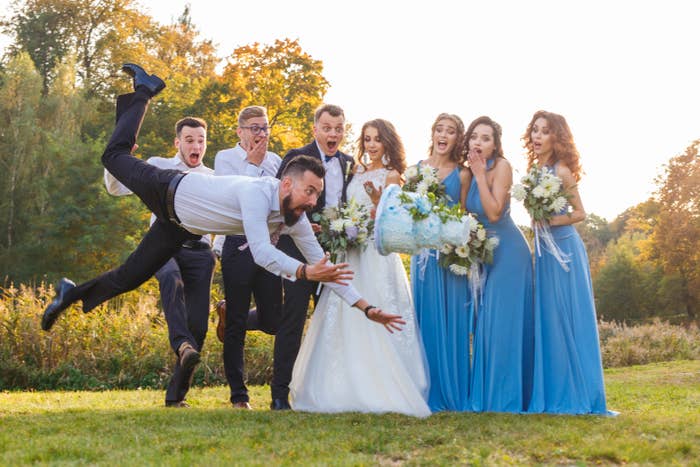 Wedding party laughing as a groomsman pretends to fall at an outdoor wedding. Bride wearing a white dress, bridesmaids in matching blue