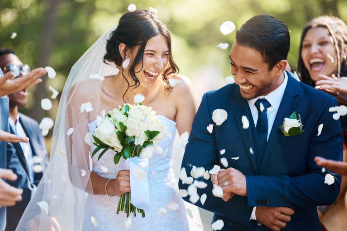 Bride and groom smiling during confetti toss at wedding with guests around. Bride holding a bouquet, groom in a suit
