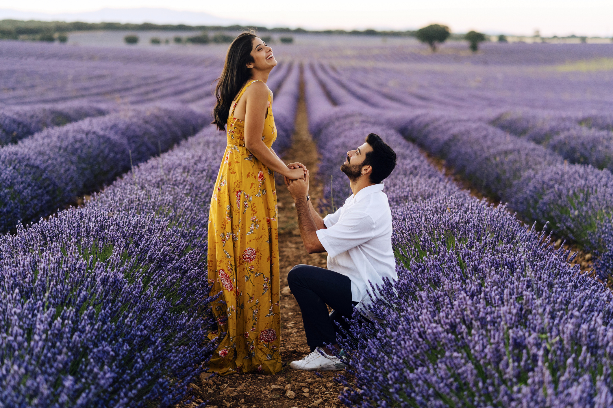Person proposing in a lavender field, partner standing, surprised; both dressed in semi-formal attire
