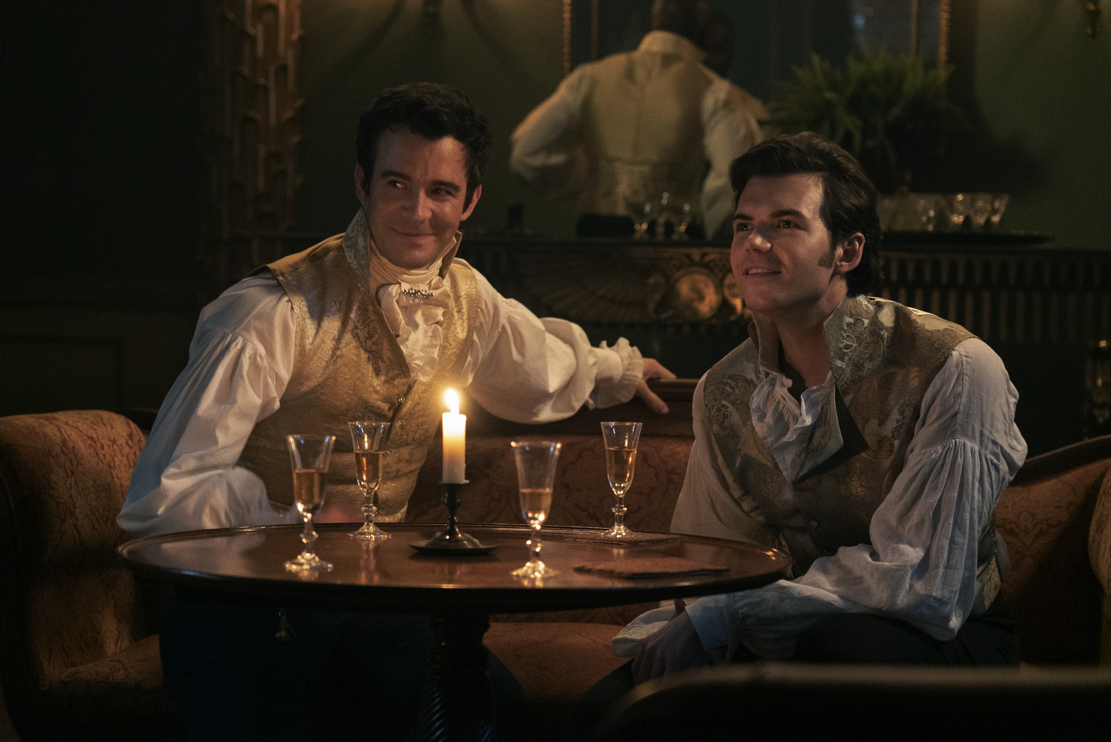 Two actors in historical costumes sitting at a table with champagne flutes and a candle, a scene from a period drama