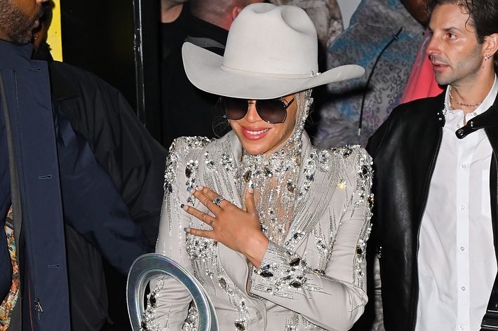 Person in embellished outfit with a wide-brimmed hat, smiling behind large sunglasses, surrounded by people