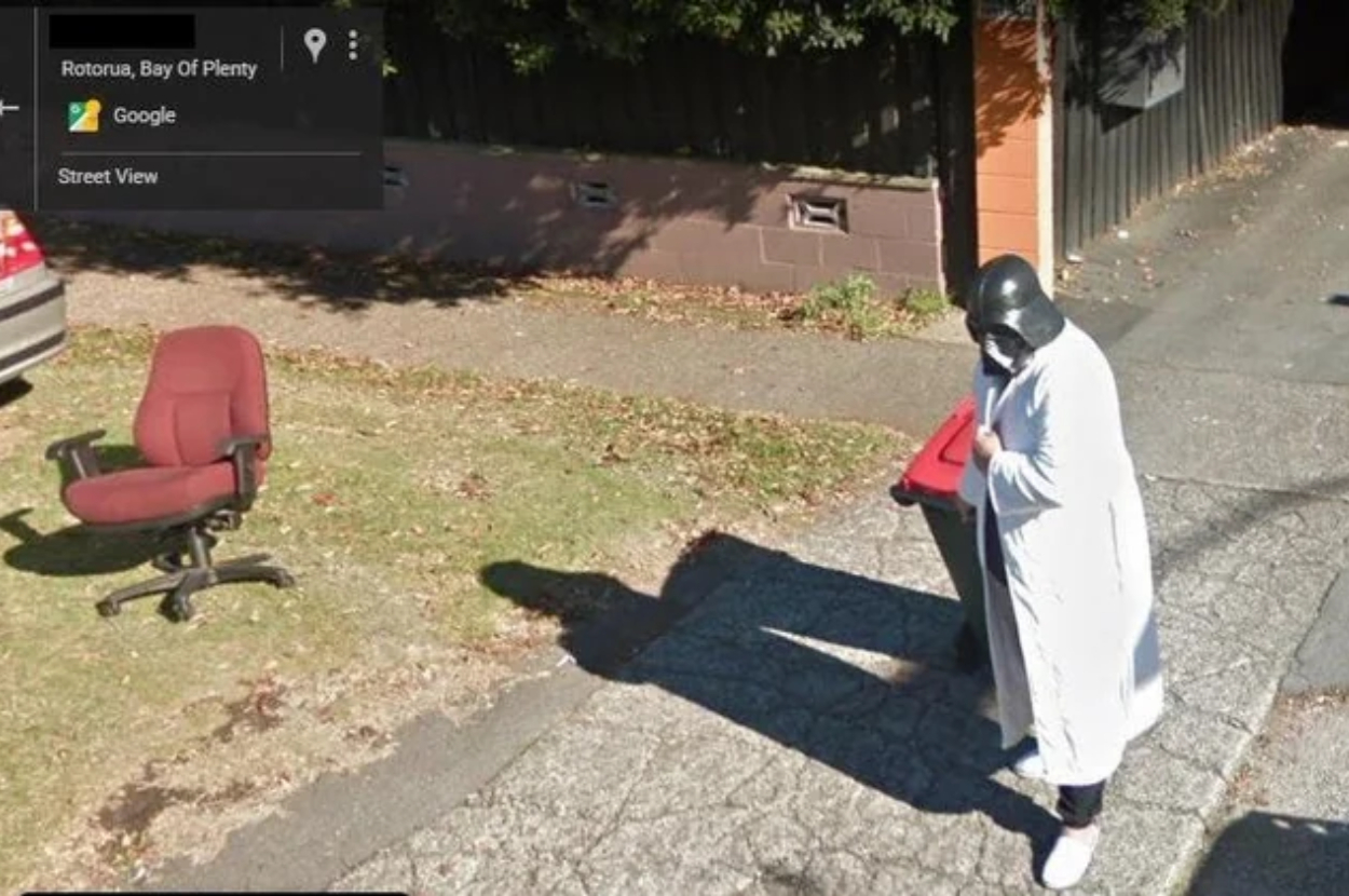 Person in a white robe and dark mask stands by an office chair on a suburban street, captured by a street view camera